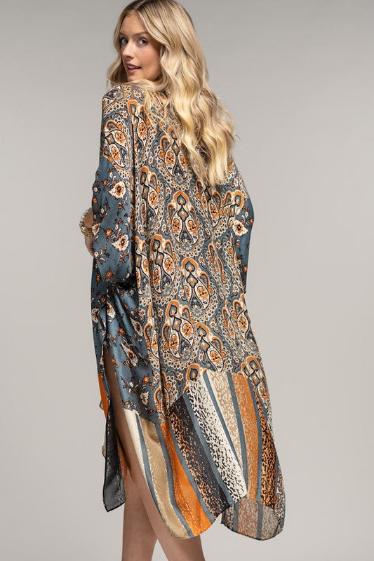woman wearing a kimono in mix of botanical and geometric patterns,  open front with side slits in shades of deep teal blue, orange and beige, back view