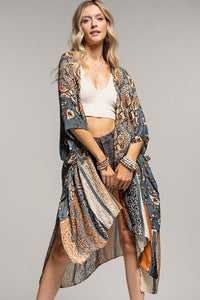 woman wearing a kimono in mix of botanical and geometric patterns,  open front with side slits in shades of deep teal blue, orange and beige