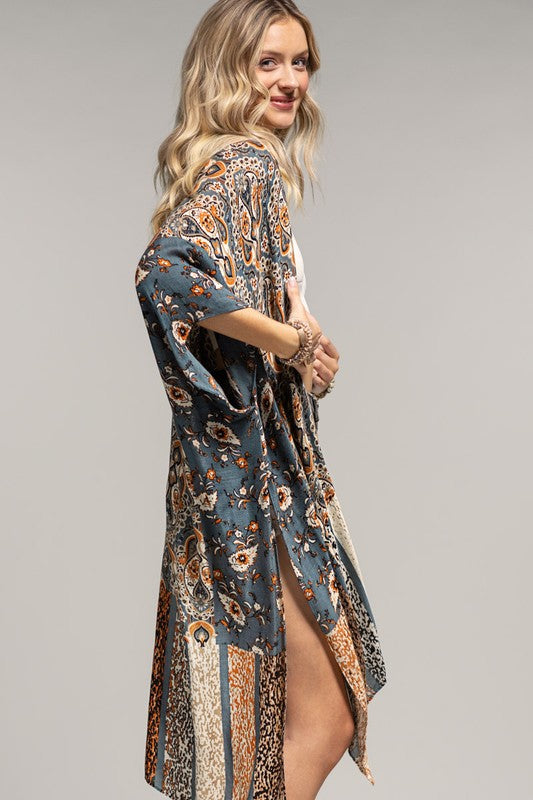 woman wearing a kimono in mix of botanical and geometric patterns,  open front with side slits in shades of deep teal blue, orange and beige, side view