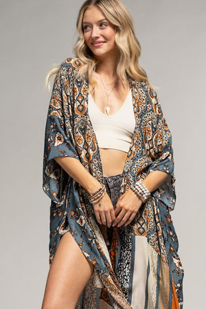woman wearing a kimono in mix of botanical and geometric patterns,  open front with side slits in shades of deep teal blue, orange and beige