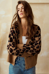 Woman wearing a leopard print  button up cardigan with wide ribbed wrists and hemline over a white top and blue jeans against a beige background