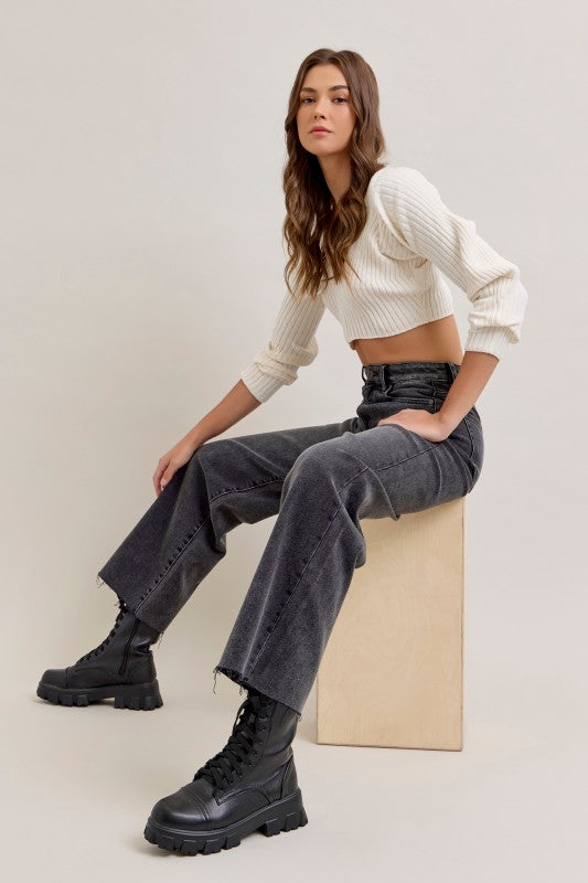Woman sitting on a beige block wearing a white cropped sweater, gray pants, and black boots.