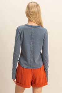 back view of woman wearing mineral washed blue t-shirt with thumb hole long sleeves and reversed stitching around collar and shoulders, exposed seam down the back