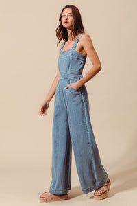 Sleeveless medium wash denim jumpsuit with pockets, back zipper closure, and wide legs