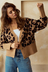 Woman wearing a leopard print  button up cardigan with wide ribbed wrists and hemline over a white top and blue jeans against a beige background